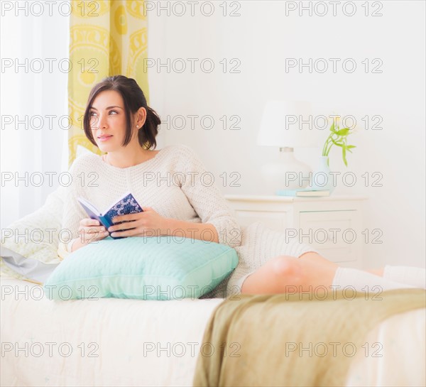 Woman leaning against pillow in bed, Holding book and looking away