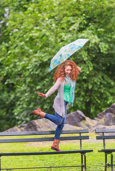 Woman with umbrella dancing on bench in park.