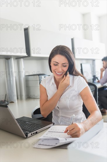 Woman talking on cell phone and doing paperwork in office, man in background.
