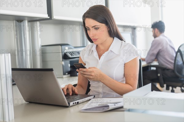 Woman working on laptop and text messaging in office, man in background.