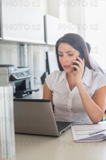 Woman working on laptop and talking on cell phone in office, man in background.