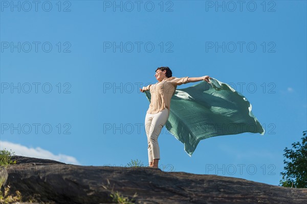 Mature woman standing on rock with arms raised.