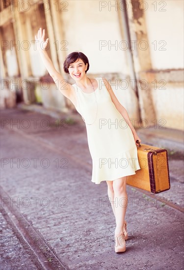 Woman in dress holding suitcase at train station, waving hand