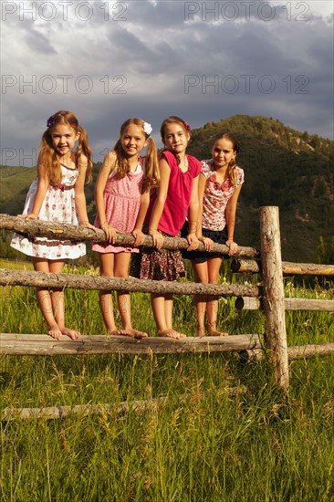 Four girlfriends (6-7, 8-9, 10-11) standing on wooden fence in grass