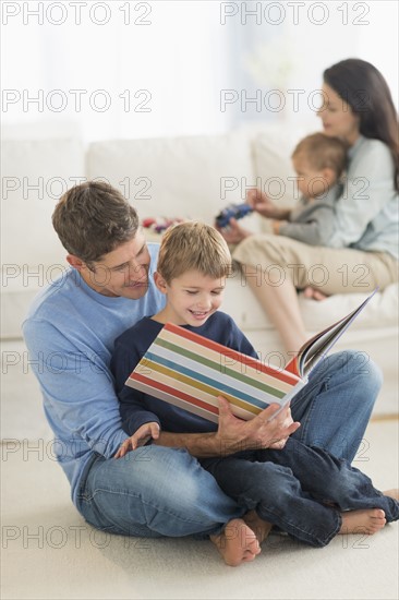 Parents with kids (12-17 months, 6-7) at home, father reading to son.