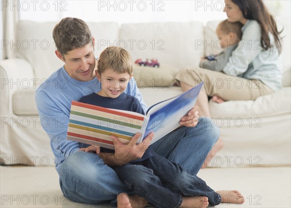 Parents with kids (12-17 months, 6-7) at home, father reading to son.