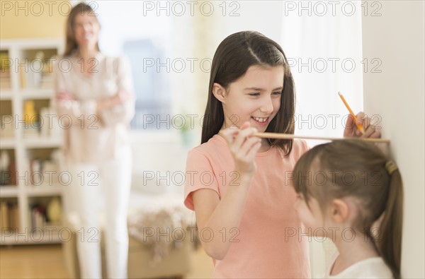 Sisters (4-5, 8-9) measuring height, mother watching.