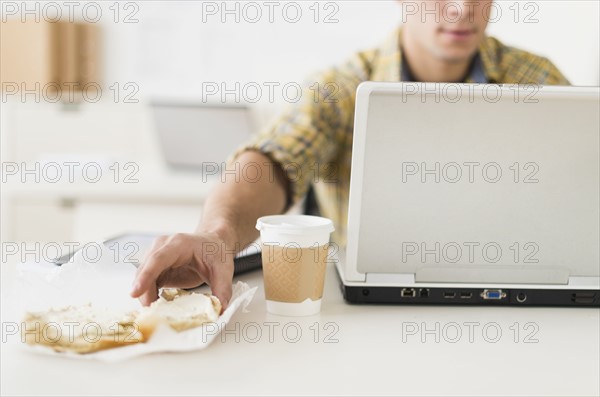 Young man eating sandwich and using laptop.