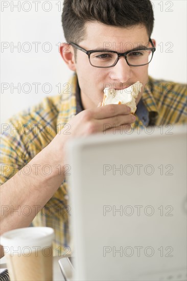 Young man eating sandwich and using laptop.