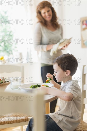 boy (6-7) eating healthy dinner, mother in background.