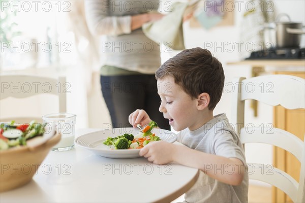 boy (6-7) eating healthy dinner, mother in background.