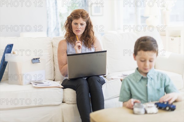 Mother using laptop, son (6-7) playing with toy cars.