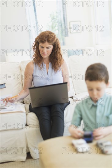 Mother using laptop, son (6-7) playing with toy cars.