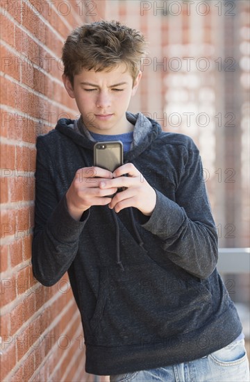 Teenage boy (16-17) leaning against brick wall texting.