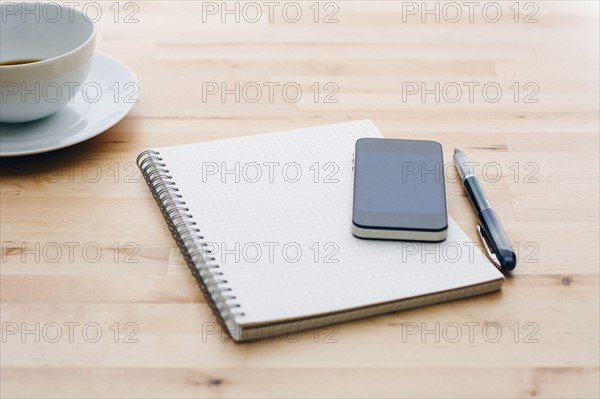 Spiral notebook, Ballpoint pen, Mobile phone and coffee cup on wooden table.
Photo : Kristin Lee