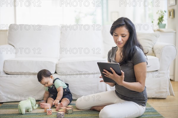 Mother using tablet pc while her son (6-11 months) playing with building blocks.