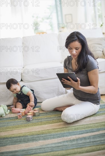 Mother using tablet pc while her son (6-11 months) playing with building blocks.