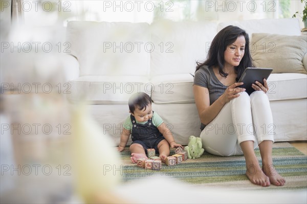 Mother using tablet pc while her son (6-11 months) playing with building blocks.