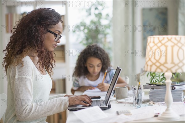 Woman using laptop at home, girl (8-9) doing homework in background.