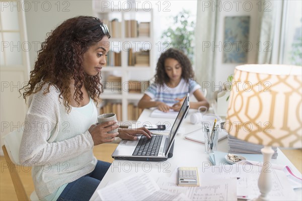 Woman using laptop at home, girl (8-9) doing homework in background.