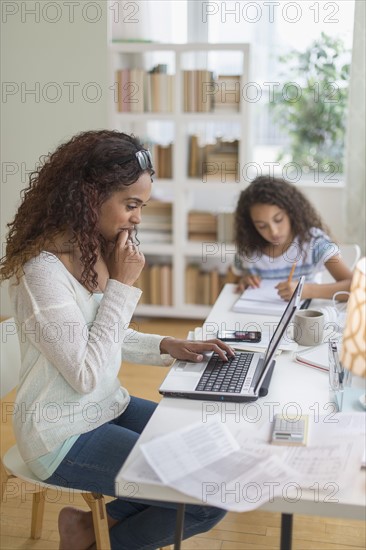Woman using laptop at home, girl (8-9) doing homework in background.