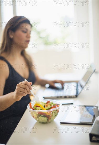 Woman eating salad and using laptop in office.