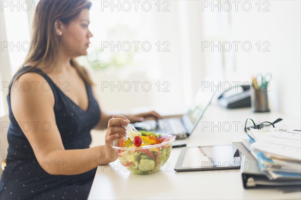 Woman eating salad and using laptop in office.