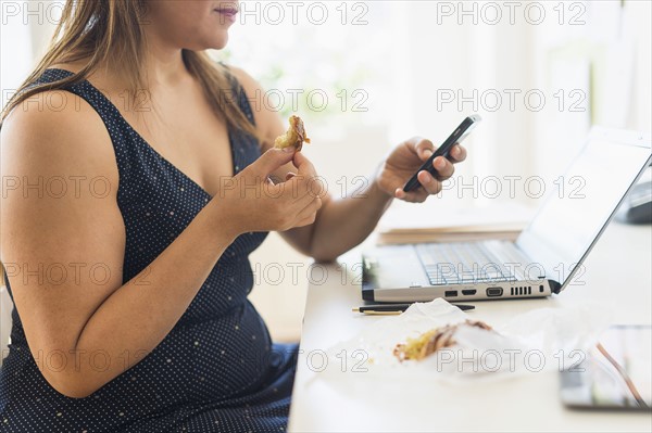 Woman eating croissant and using mobile phone in office.