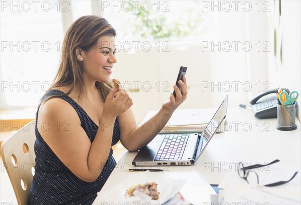 Woman eating croissant and using mobile phone in office.