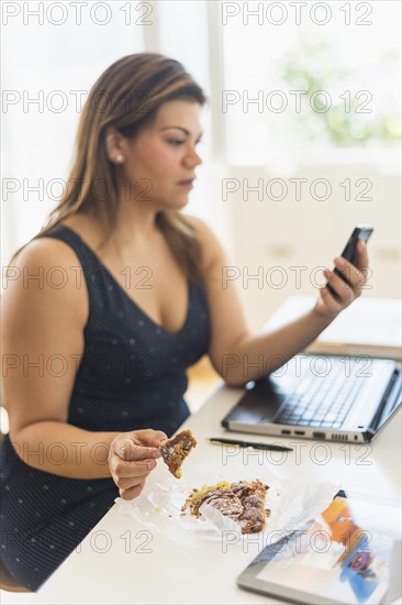 Woman eating croissant and using mobile phone in office.