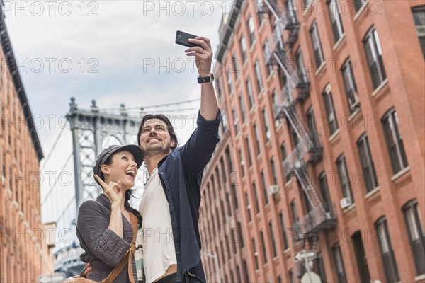 Couple taking selfie on street, Brooklyn Bridge in background. Brooklyn, New York.