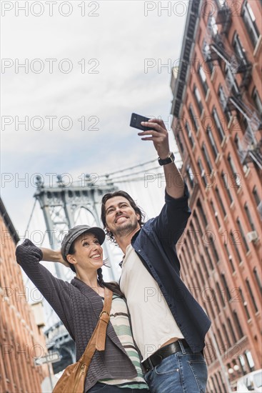 Couple taking selfie on street, Brooklyn Bridge in background. Brooklyn, New York.