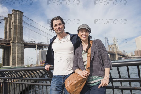 Couple leaning against railing and looking at camera. Brooklyn, New York.