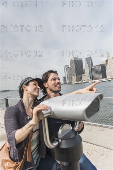 Happy couple sitting by coin-operated binoculars. Brooklyn, New York.