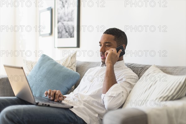 Man sitting in living room, using laptop and cell phone.