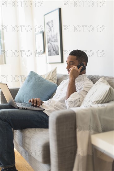 Man sitting in living room, using laptop and cell phone.