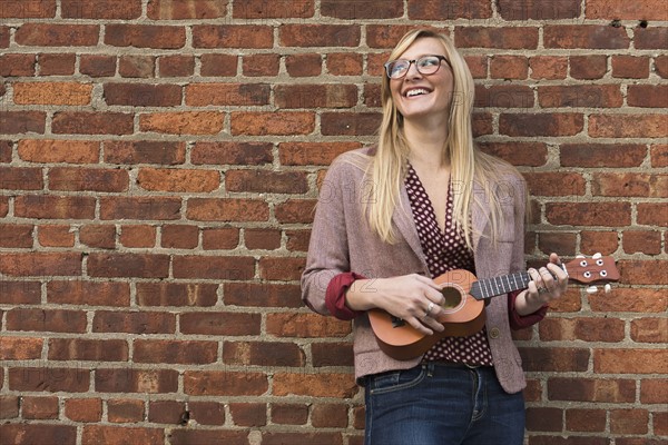Woman standing against brick wall and playing ukulele.