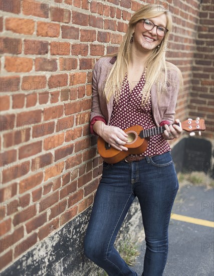 Woman standing against brick wall and playing ukulele.