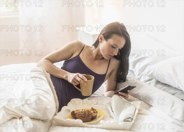 Woman having breakfast in bed, using mobile phone.