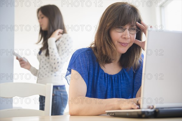 Mother using laptop, daughter (14-15) using mobile phone in background