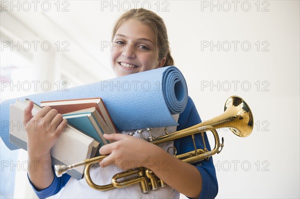 Portrait of teenage girl (12-13) holding rolled-up exercise mat, books, and trumpet