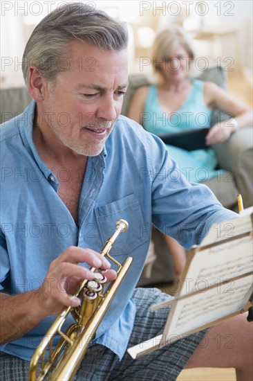 Portrait of senior man holding trumpet looking at music stand with woman on sofa in background