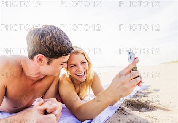 Portrait of young couple lying on blanket on beach, using smart phone