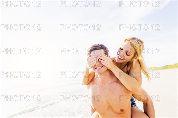 Girlfriend riding piggyback on beach, closing boyfriend's eyes with palms