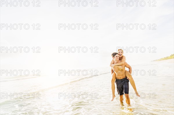 Boyfriend walking in surf carrying girlfriend piggyback