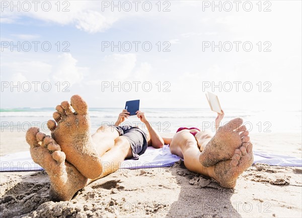 Close-up of sand-covered feet of young couple lying on blanket on beach reading