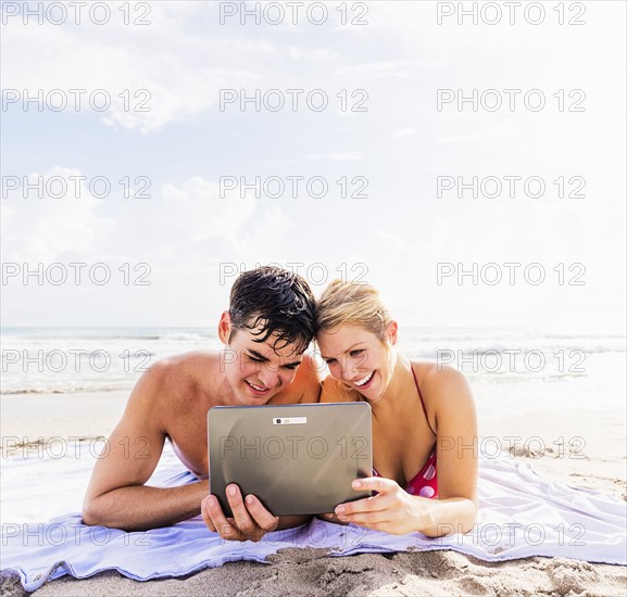 Portrait of young couple lying on blanket on beach side by side, using digital tablet