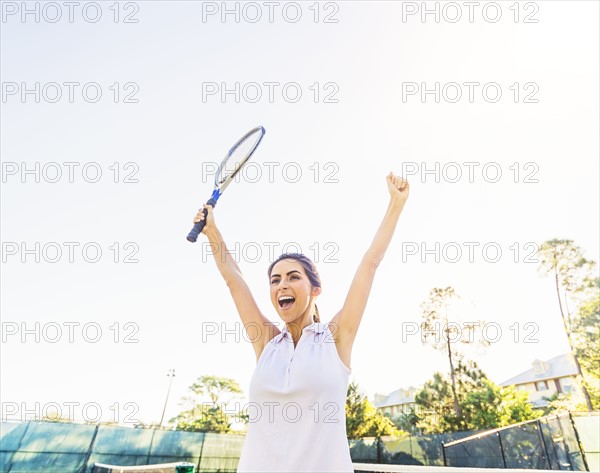 Portrait of young woman holding tennis racket, raising arms in celebration
