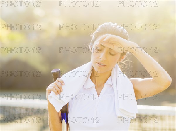 Portrait of young woman with towel and tennis racket wiping forehead with back of hand