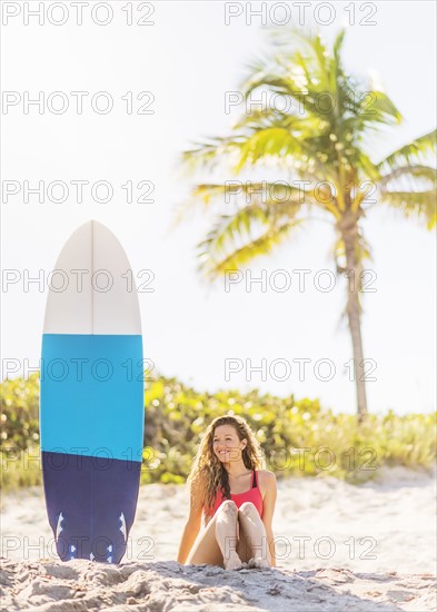 Portrait of young woman sitting near surfboard on beach